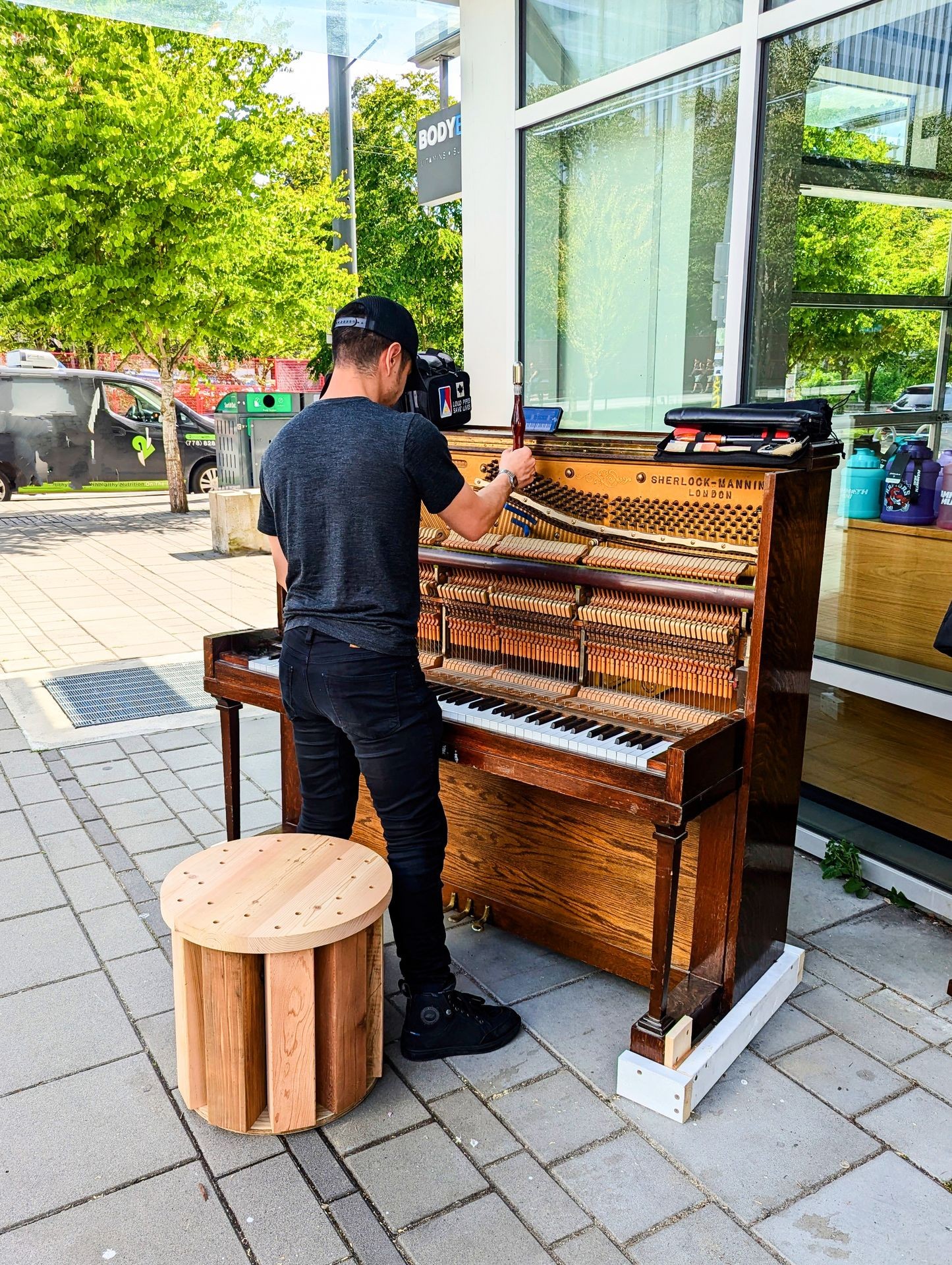 Person tuning an upright piano outside on the sidewalk, with a tree and building in the background.