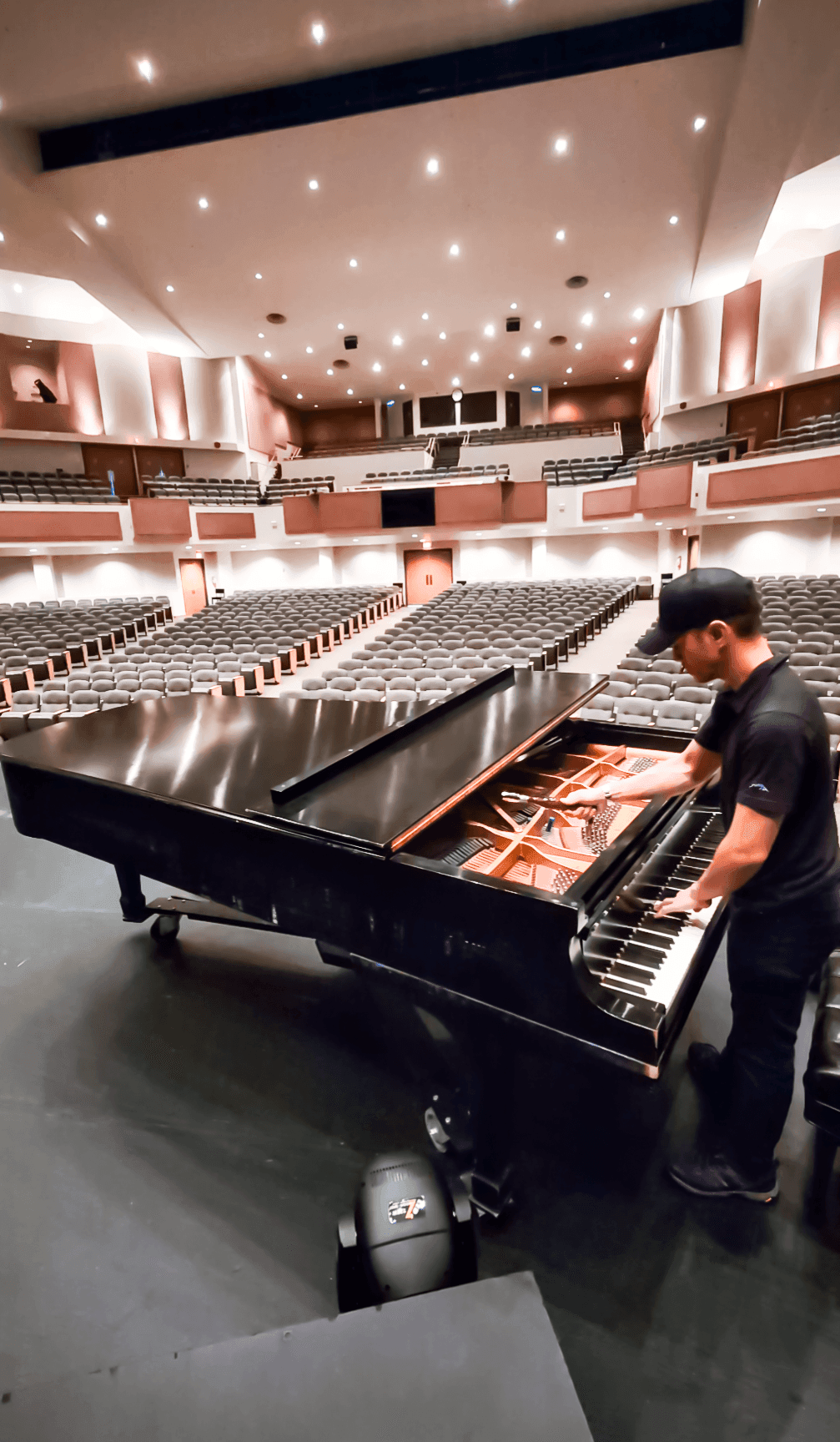 Person tuning a grand piano on an empty concert hall stage with rows of seats and illuminated ceiling.